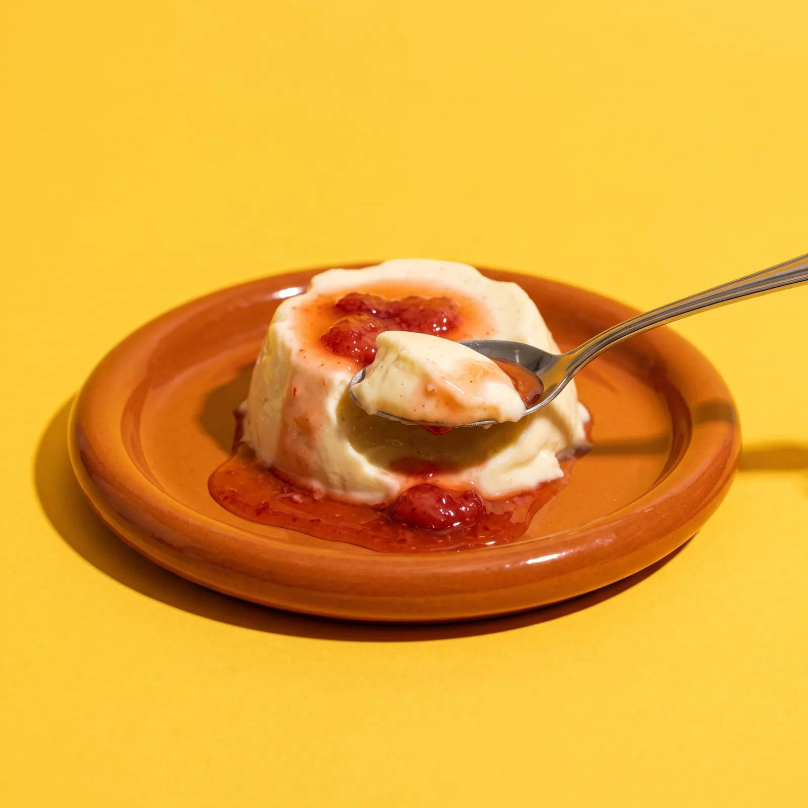 Panna cotta con salsa de frutilla en plato de greda, una cuchara levantando una porción, sobre fondo amarillo