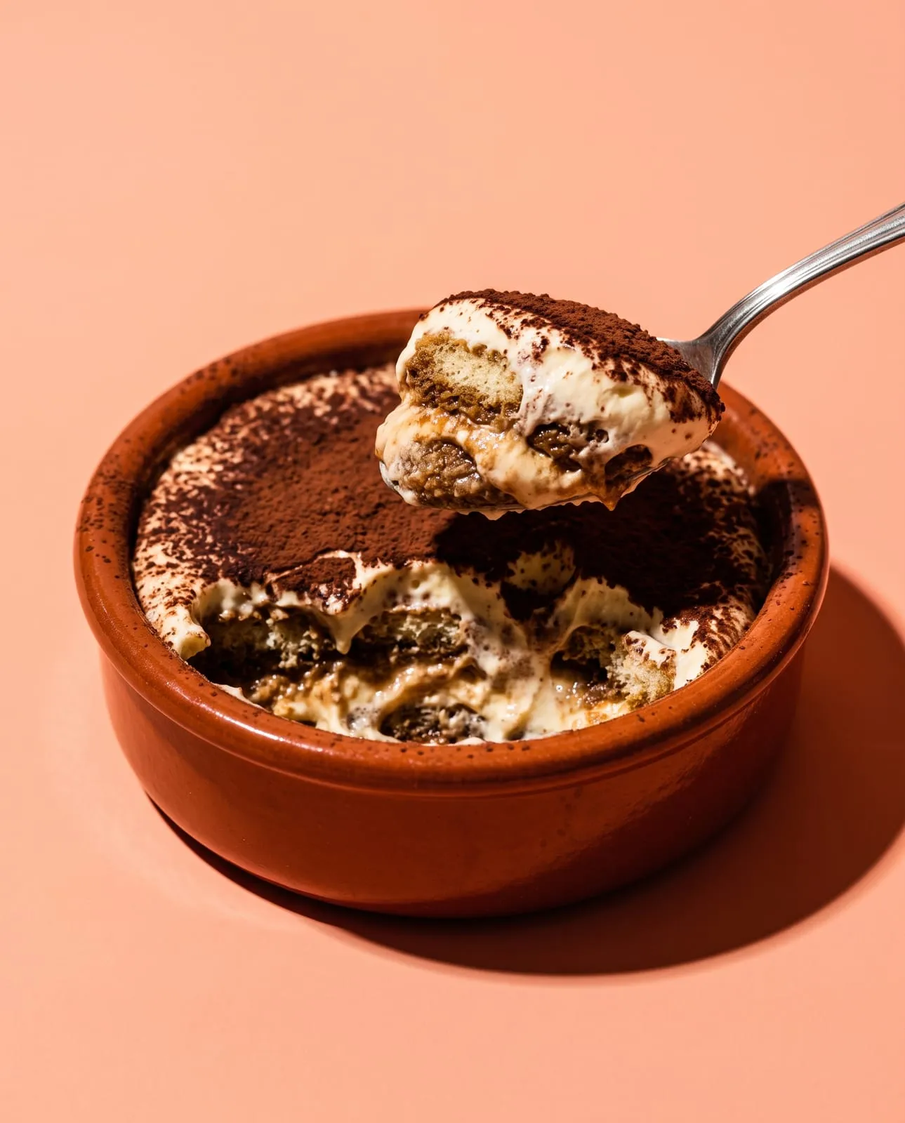 Tiramisù con cacao en cazuela de greda, una cuchara levantando una porción, sobre fondo durazno
